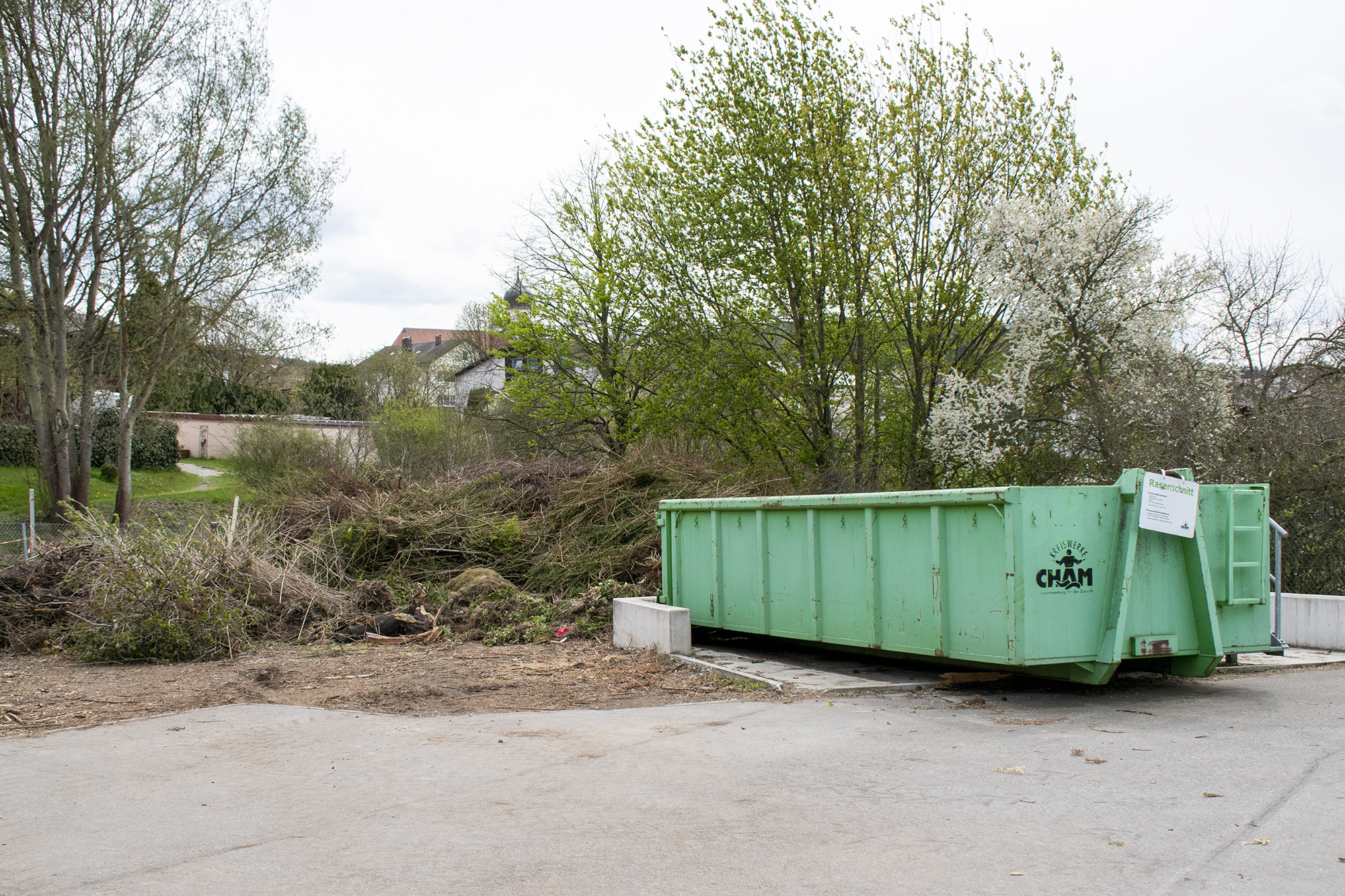 Grüner Container für Rasenschnitt mit dahinter liegendem Schnittholz